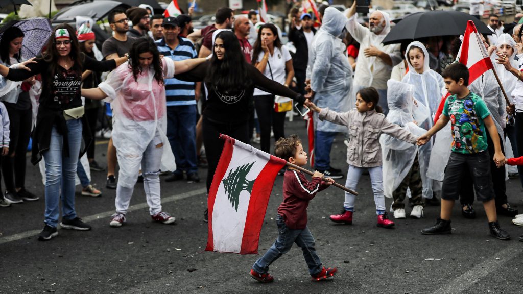 A young boy walks with a Lebanese flag past protesters, some wearing ponchos, gathering during a demonstration on the eighth day of protest against tax increases and official corruption, in Zouk Mosbeh, north of the capital Beirut, on October 24, 2019. - The almost one-week-old massive street protests in Lebanon, sparked by a tax on messaging services such as WhatsApp, have morphed into a united condemnation of a political system seen as corrupt and beyond repair. (Photo by JOSEPH EID / AFP)